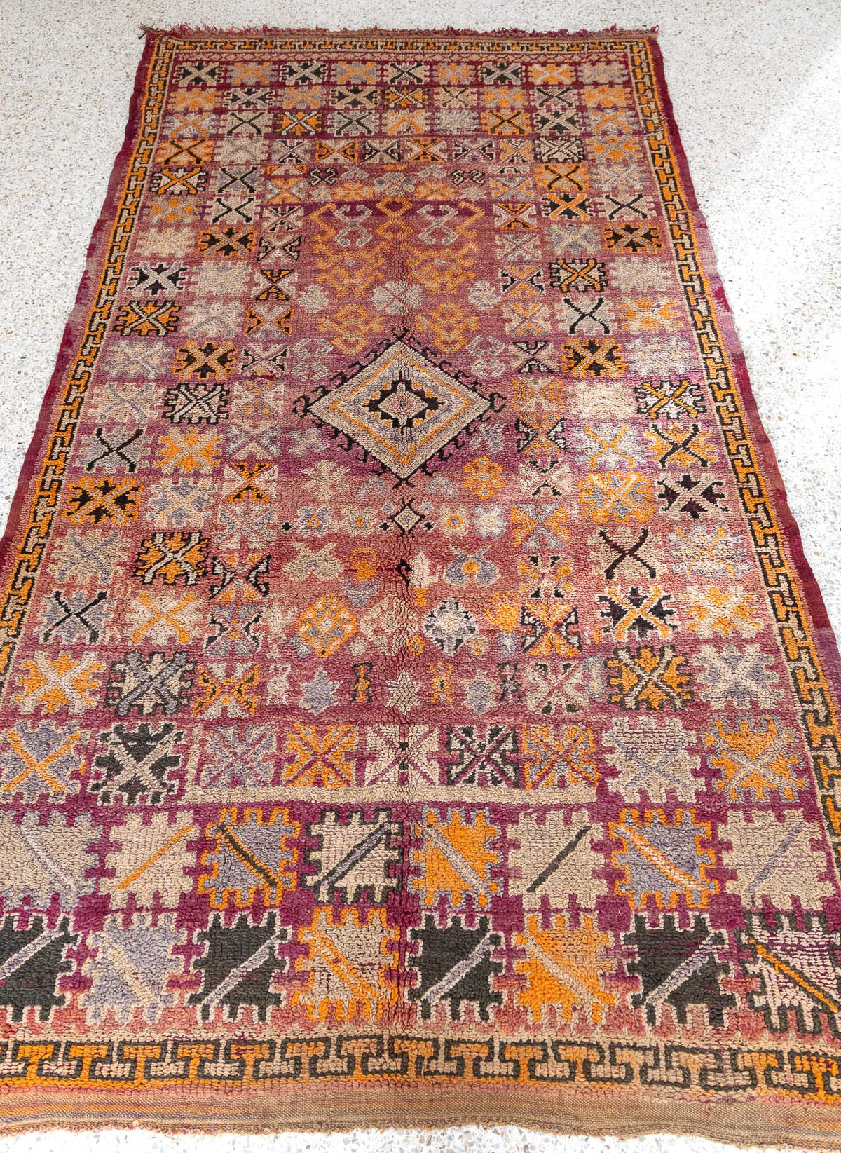 The carpet is a real feast for the eyes, with a mixture of colors and Berber signs in a sophisticated and elegant composition. The design is dense and intricate, with a symmetrical pattern that has variations in the organization of the composition. The rug features a central diamond surrounded by a rectangular frame with a different pattern, which adds an interesting visual element to the composition.  faded color palette from rosy background to yellowish beige, and dark.
